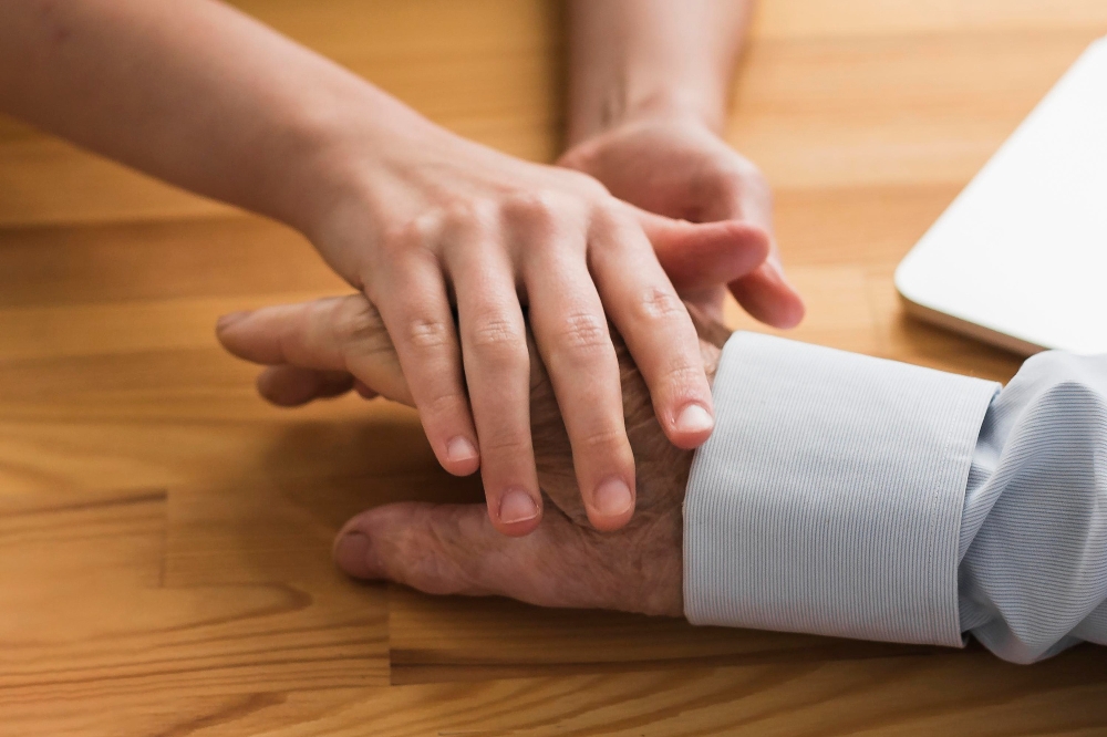 Caregiver holds elderly patient's hand on wooden table surface.